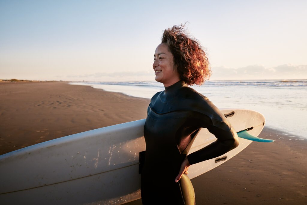 side view portrait of a woman standing in the surf with a surfboard under her arm.