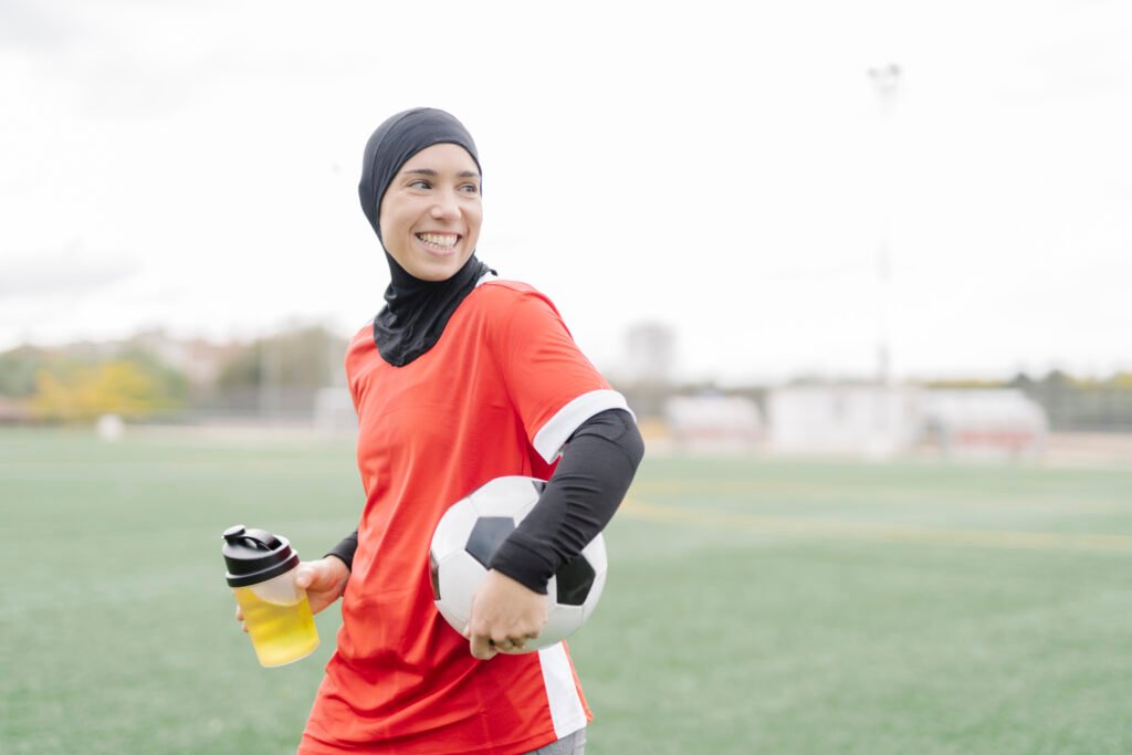 smiling sporty arabic woman drinking something to refresh herself after soccer training on the field.