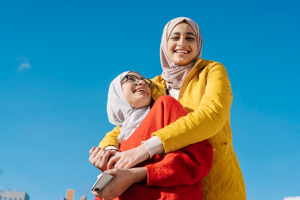 two muslim female friends hugging and smiling while posing outdoors against a clear blue sky.