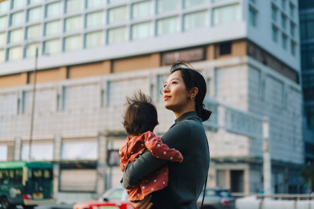 young asian mother holding her little daughter in her arms while walking down the city street, with modern city skyscrapers and busy traffic in background. family with kids lifestyle
